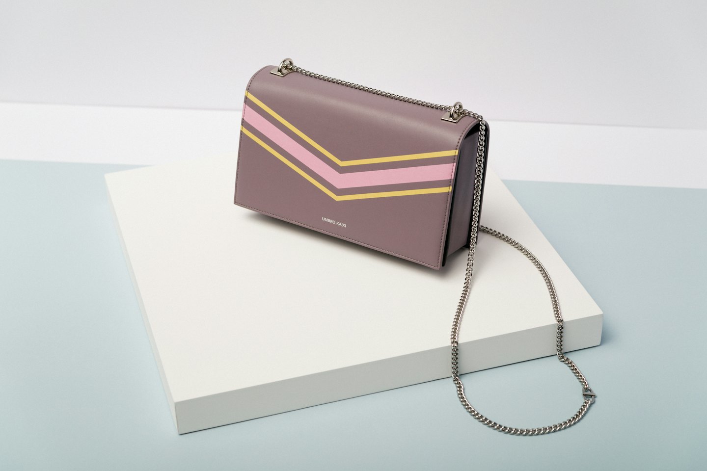 Mauve shoulder bag with pink and yellow stripes on a white plinth.